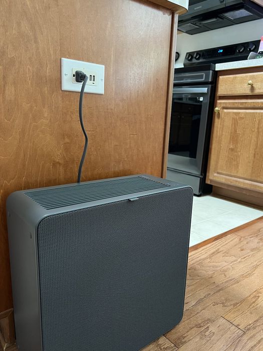 A black air purifier plugged into a kitchen outlet, positioned next to wooden cabinets and a stove.
