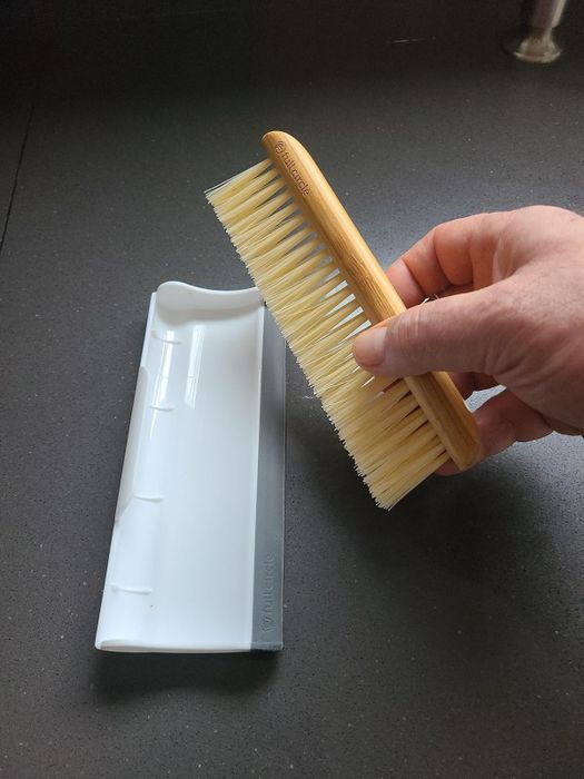 A hand lifts a wooden brush above a white and gray plastic cleaning tray, showcasing a set of scrubbing tools.