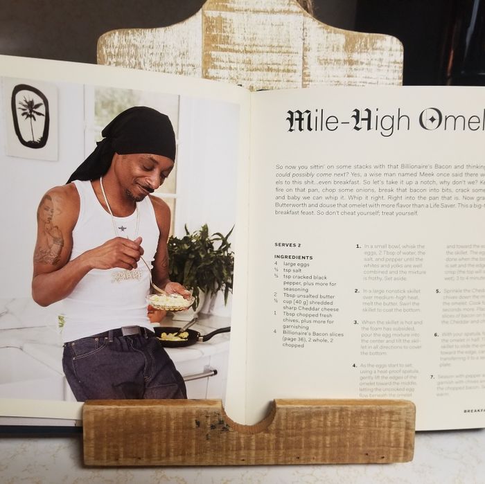 A photo shows Snoop Dogg in a kitchen, rocking a white tank top and black headscarf, as he features in a cookbook with a recipe for a Mile-High Omelet resting in the stand.