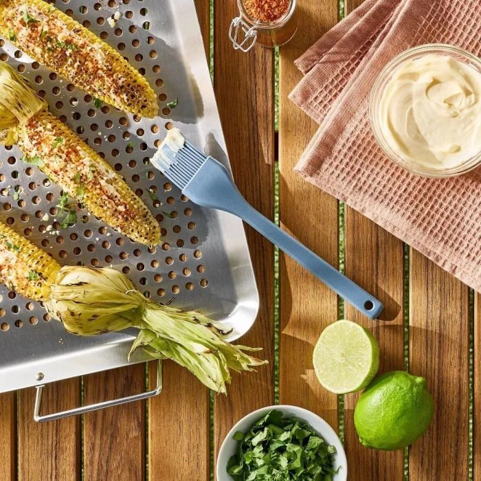 Grilled corn sprinkled with spices and herbs, resting on a metal tray, accompanied by a basting brush, lime halves, bowls of herbs and sauce, and a spice jar on a wooden table.