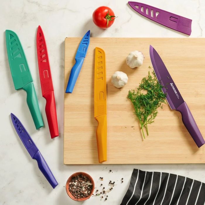 A set of six kitchen knives, each equipped with blade guards, displayed on a counter beside a chopping board with fresh herbs and garlic ready for meal prep.