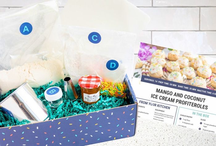 A kit containing neatly organized baking supplies, colorful sprinkles, a jar of jam, a piping tip, and a recipe card for Mango and Coconut Ice Cream Profiteroles.
