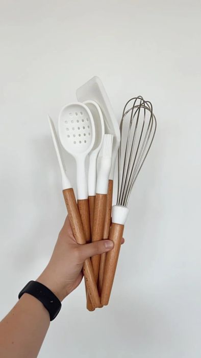 A hand holding wooden-handled kitchen utensils, including a spatula, spoon, and whisk.