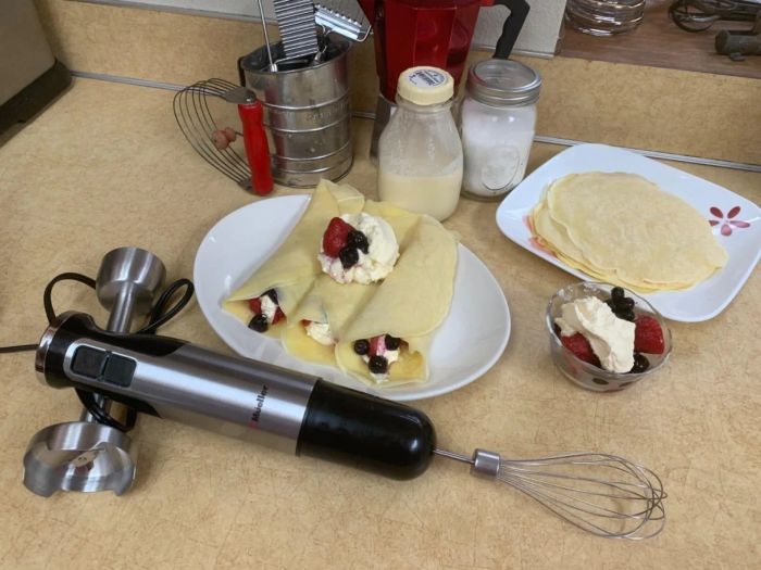 A kitchen counter topped with crepes filled with cream and berries, a jug of milk, and an immersion blender accompanied by various attachments.