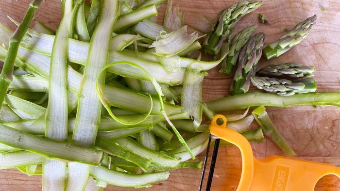 Asparagus shavings and tips placed on a wooden cutting board with a vegetable peeler.