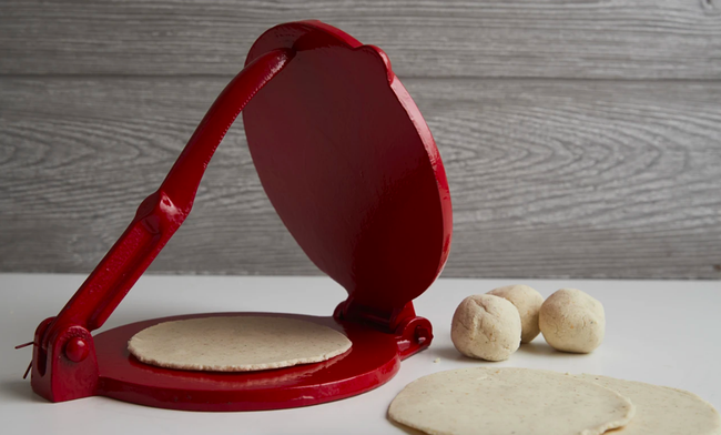 A bright red tortilla press surrounded by balls of dough and neatly flattened tortillas