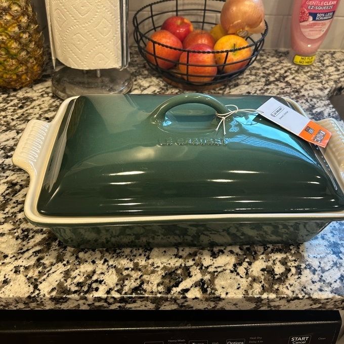 A green ceramic baking dish with a lid on a kitchen counter, tagged with a price, surrounded by fresh fruit and a roll of paper towels.