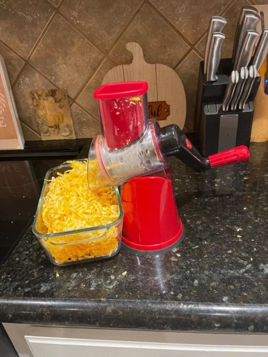 A countertop with a red rotary cheese grater filled with shredded cheese, accompanied by a glass container.
