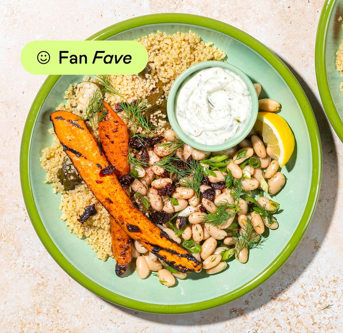 A plate of couscous served with grilled carrots, beans, fresh herbs, and a lemon wedge, topped with a generous spoonful of white sauce.