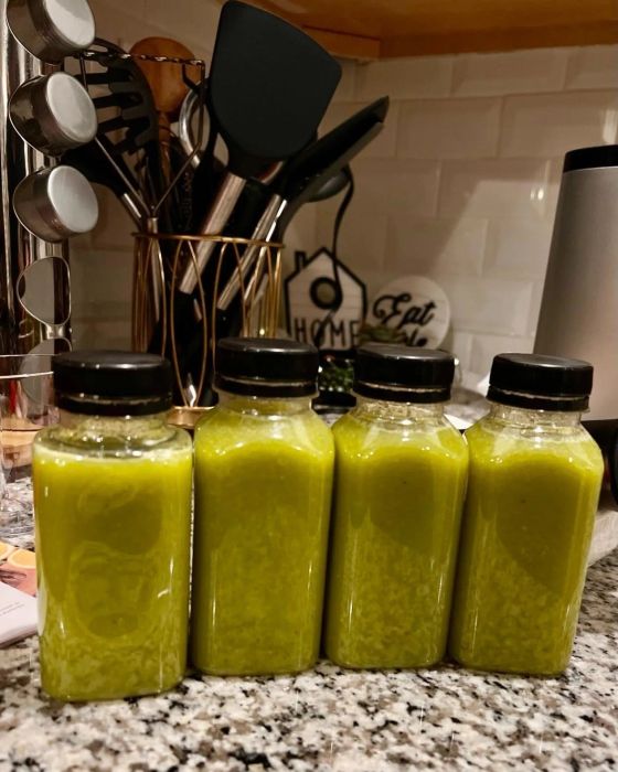 Four bottles of freshly made green juice placed on a kitchen counter with utensils visible in the background