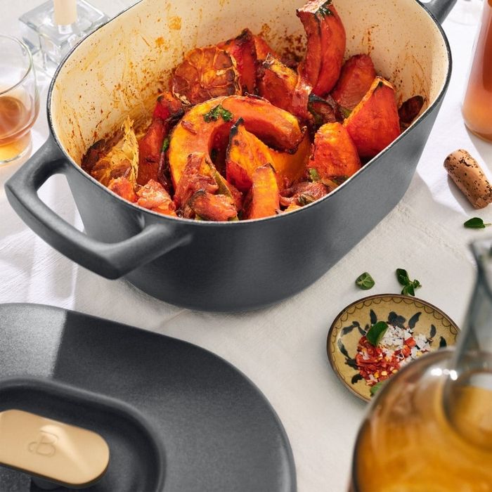 A cast iron Dutch oven filled with roasted squash and herbs, alongside a bottle of olive oil and a small plate of spices on a white tablecloth
