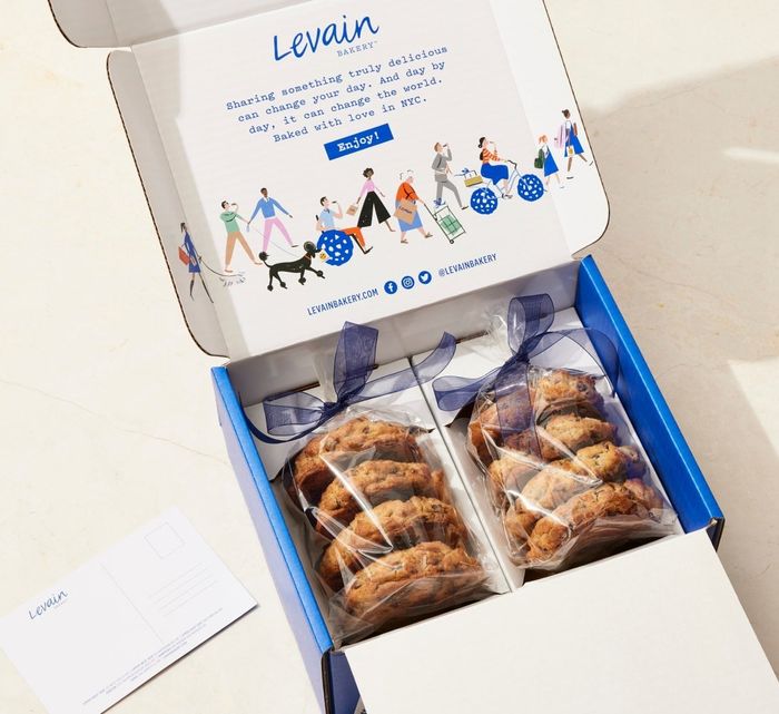 Inside a Levain Bakery box, two packs of freshly baked cookies are tied with ribbon. The box proudly displays the Levain Bakery logo, along with a note about their delicious treats.