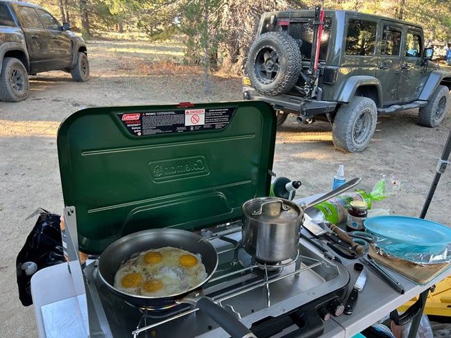 A reviewer’s camp stove set up at a campsite, with a pot cooking on one burner and a pan frying eggs on the other