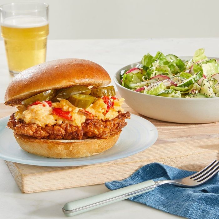 A crispy chicken sandwich loaded with pimento cheese and pickles, served alongside a green salad and a refreshing glass of beer