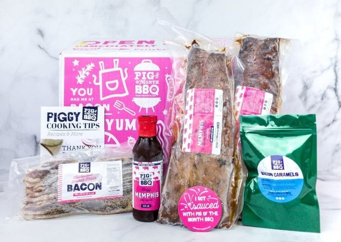 A cardboard box containing a bottle of BBQ sauce, uncooked bacon, two cuts of meat, a bag of bacon caramels, and a cooking tips pamphlet.