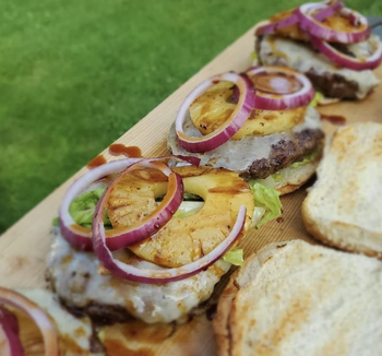 A wooden platter with open-faced burgers topped with pineapple, red onion slices, and lettuce, next to golden, toasted buns