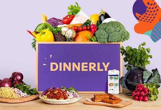 A Dinnerly meal kit filled with fresh vegetables, with a prepared meal on a plate in the foreground, alongside another dish and a smartphone displaying a recipe