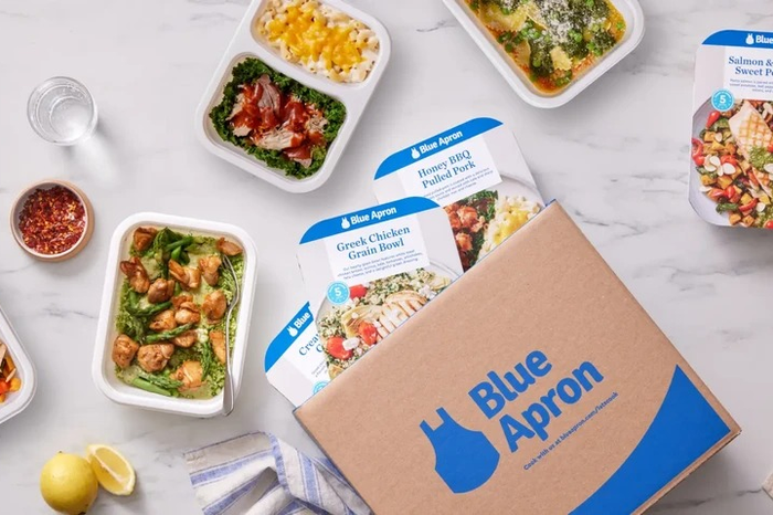Blue Apron meal kits, featuring Honey BBQ Pulled Pork, Greek Chicken Grain Bowl, and more, displayed next to a Blue Apron delivery box