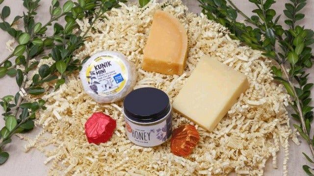 A delightful assortment of cheeses, a small jar of honey, and decorative items placed on a bed of shredded paper, all surrounded by fresh greenery.