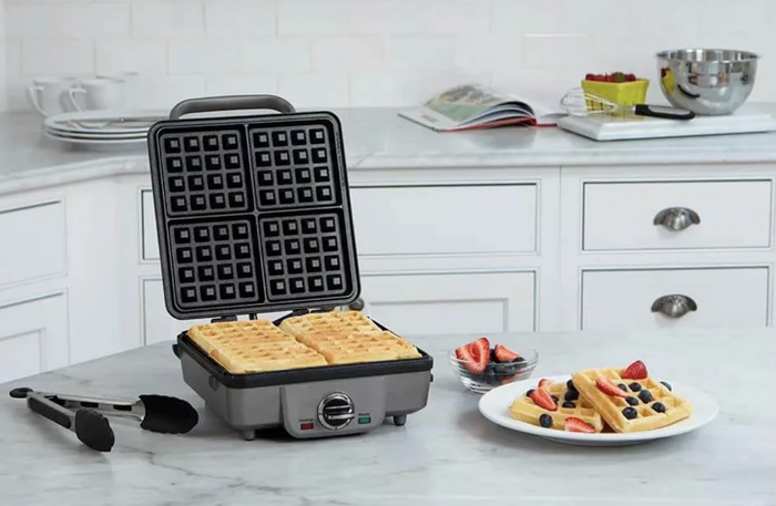 An open waffle iron with freshly cooked waffles on a kitchen counter, accompanied by a plate of waffles topped with berries and a bowl of strawberries in the background