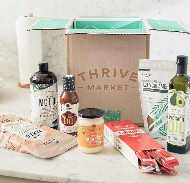 An open Thrive Market box surrounded by a variety of grocery items like BBQ sauce, jerky sticks, and avocado oil