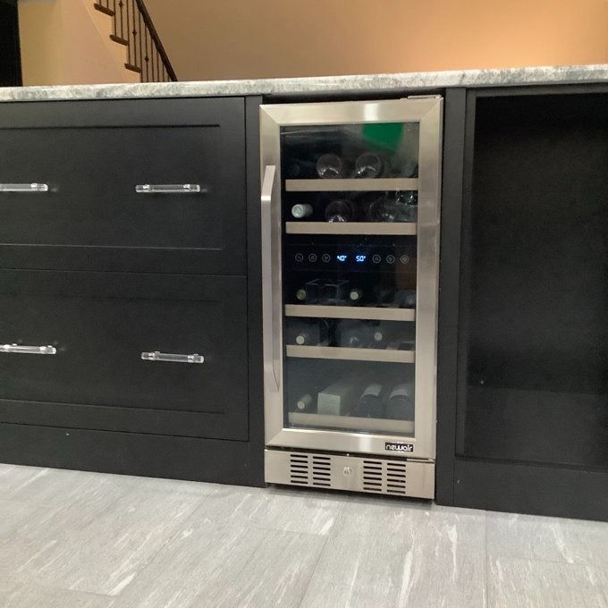 A built-in wine cooler with a glass door, partially filled with bottles, seamlessly integrated into sleek black cabinetry beneath the countertop.