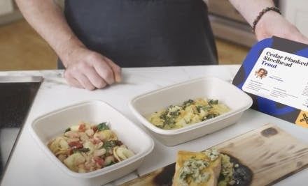 A person prepares a dish from a Cedar Planked Steelhead Trout meal kit, displaying pasta dishes and the kit's packaging on a kitchen counter.