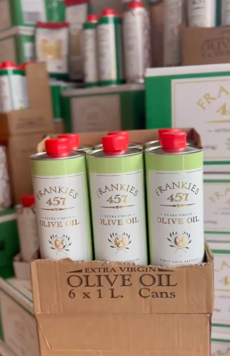 Frankies 457 Extra Virgin Olive Oil in tall cans, neatly displayed in a cardboard box with many more stacked behind it.