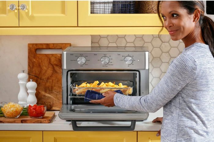 A model pulls a basket of French fries from the toaster oven, which is equipped with a wired basket for easy air frying