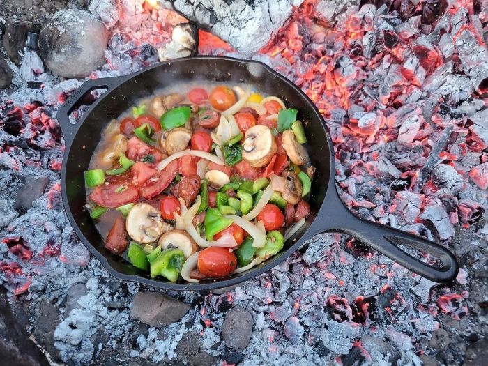 A cast iron skillet with mixed vegetables cooking over hot coals.
