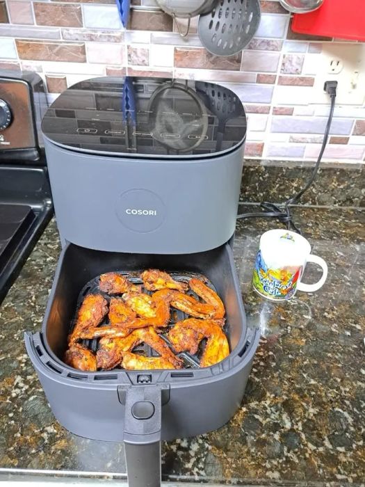 Cosori air fryer placed on the kitchen counter, filled with cooked chicken wings, alongside a mug