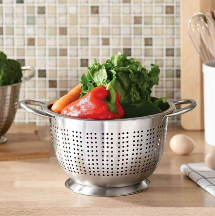 A stainless steel colander brimming with fresh produce on a kitchen counter, described as food-grade, dishwasher-safe, freezer-safe, and resistant to rust and dents