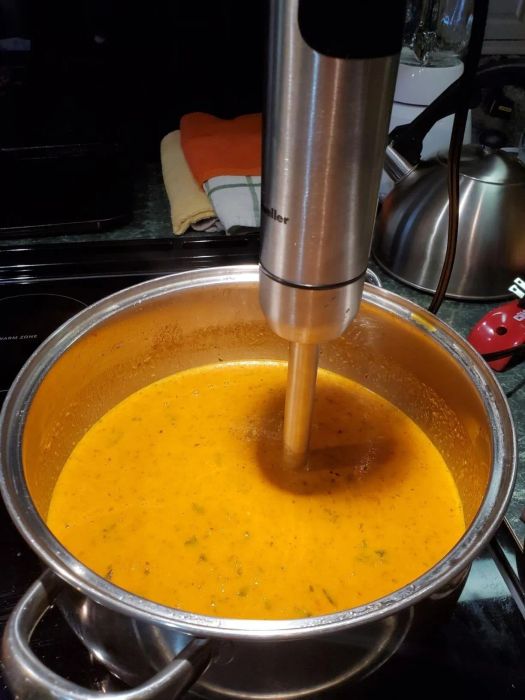 An immersion blender in action, blending soup in a pot, with a kitchen background.