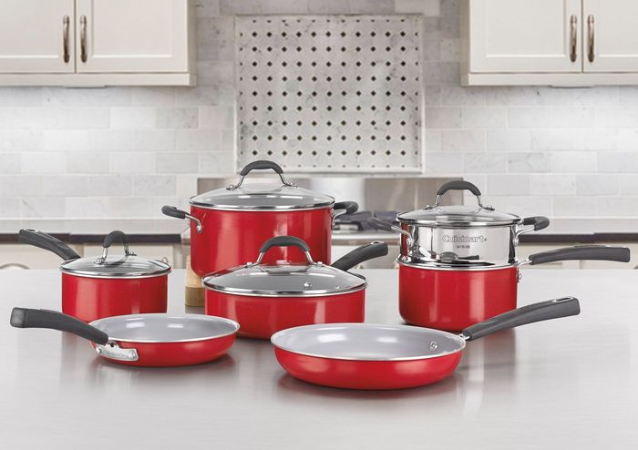 A set of red ceramic cookware, including pots, pans, and storage containers, neatly displayed on a kitchen counter with white cabinets and a backsplash in the background