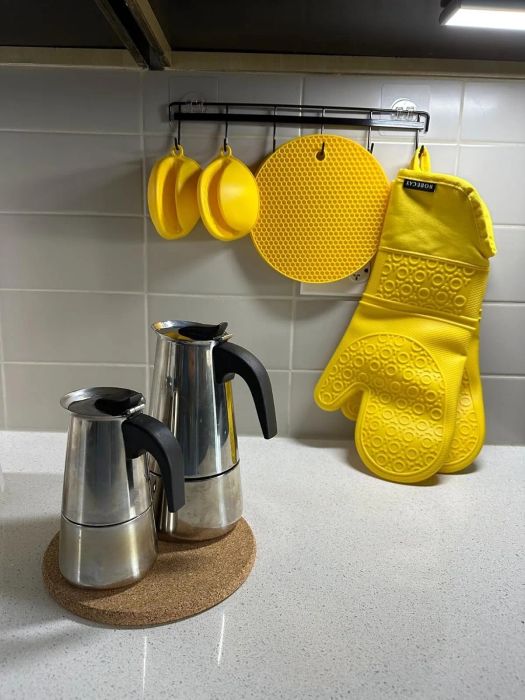 Two shiny stainless steel coffee pots resting on a cork trivet, with a pair of yellow oven mitts hanging nearby on a kitchen rack.