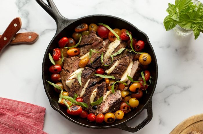 A skillet showcasing grilled steak, cherry tomatoes, and herbs on a marble surface, accompanied by a knife, napkin, and basil