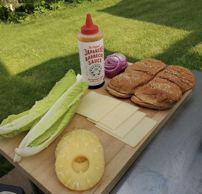 A spread of ingredients for preparing burgers: romaine lettuce, a pineapple ring, cheese slices, red onion, sesame seed buns, and Japanese barbecue sauce