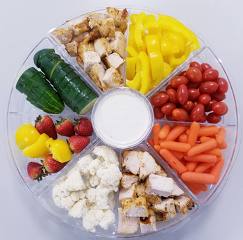 A platter loaded with sliced chicken, yellow bell peppers, cherry tomatoes, baby carrots, cauliflower, strawberries, and cucumber slices, all accompanied by a dip in the middle