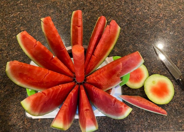 A radial arrangement of sliced watermelon on a kitchen counter, featuring the cylindrical melon core in the center, accompanied by a small knife and two round end pieces.