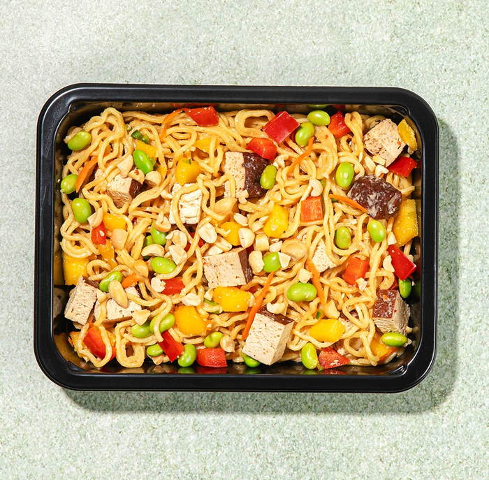 A black tray filled with a noodle dish featuring tofu cubes, edamame, bell peppers, and an assortment of other vegetables.