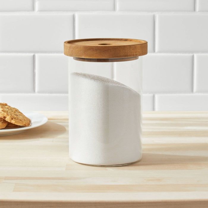 A glass canister with a wooden lid filled with sugar on a kitchen counter, placed next to a plate of cookies