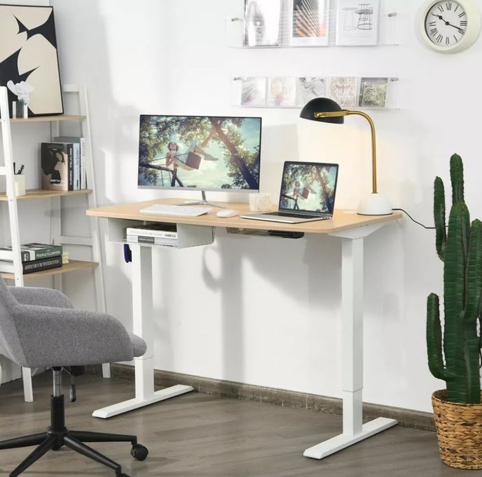 A modern home office setup with a standing desk, dual monitors, and a laptop. Nearby, a gray chair and a cactus add some life. The minimalist decor on the shelves and walls completes the sleek look.