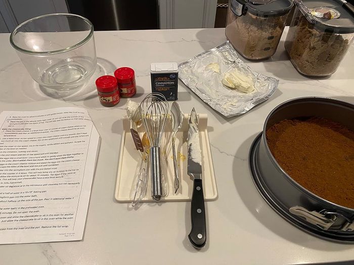 A kitchen counter setup featuring a recipe, baking ingredients, and tools like a mixer and springform pan, ready for baking
