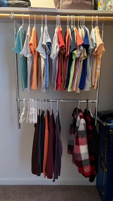 A closet neatly organized with folded tops on the upper rack and jackets hanging below, with empty hangers on the side