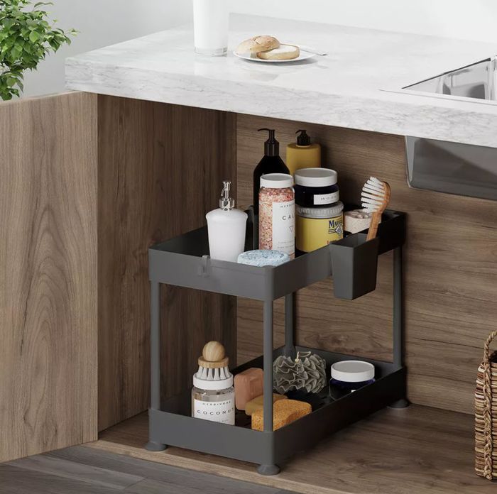 An under-sink organizer with neatly arranged toiletries, soaps, and sponges on two shelves next to a kitchen counter.