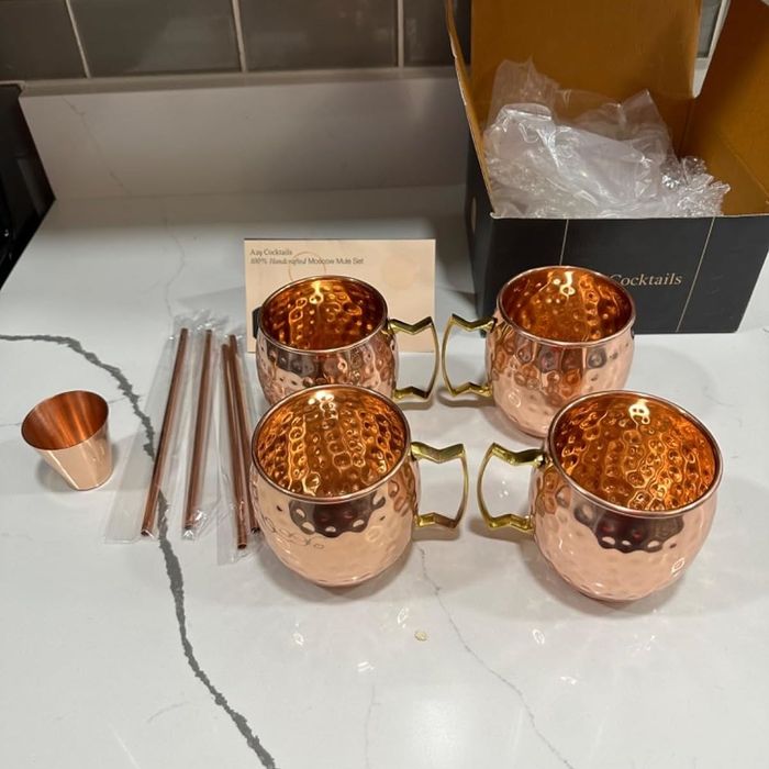 Four copper mugs with handles, a measuring cup, and straws arranged on a kitchen counter, likely for preparing Moscow Mule cocktails