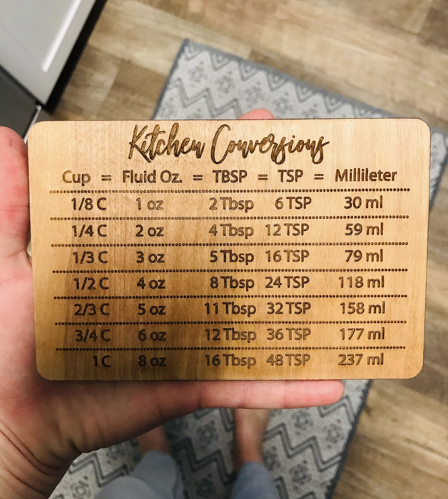 A person holding a wooden kitchen conversion chart displaying measurements for cups, fluid ounces, tablespoons, teaspoons, and milliliters