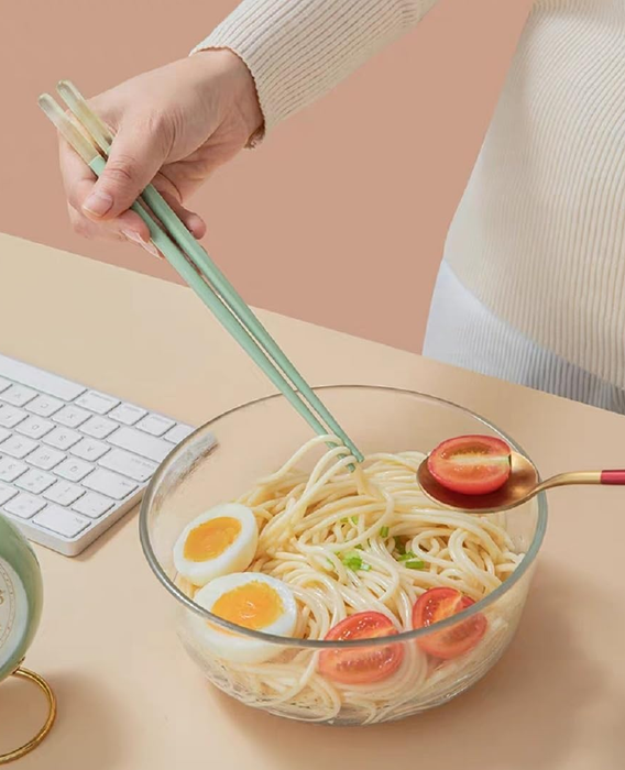 A person using chopsticks to grab noodles from a glass bowl containing eggs and tomatoes, placed near a keyboard