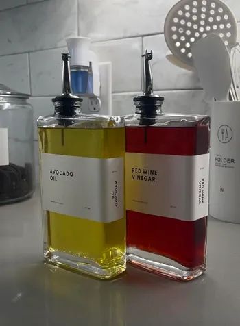 Two square glass bottles with pour spouts, labeled “Avocado Oil” and “Red Wine Vinegar,” sitting on a kitchen counter
