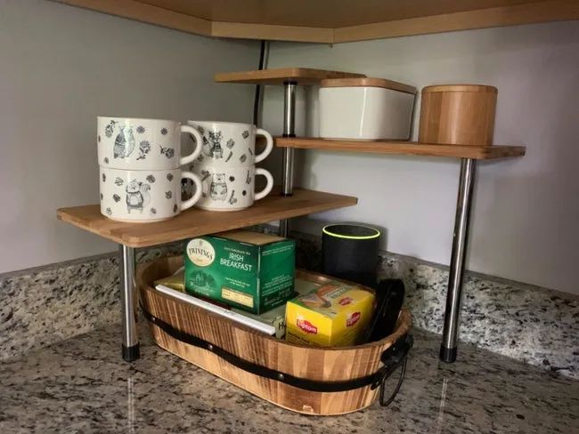A reviewer’s photo featuring the three-tier corner shelf with wood tops and silver legs, positioned in the corner of a counter, holding mugs and various items on both the top and bottom shelves.